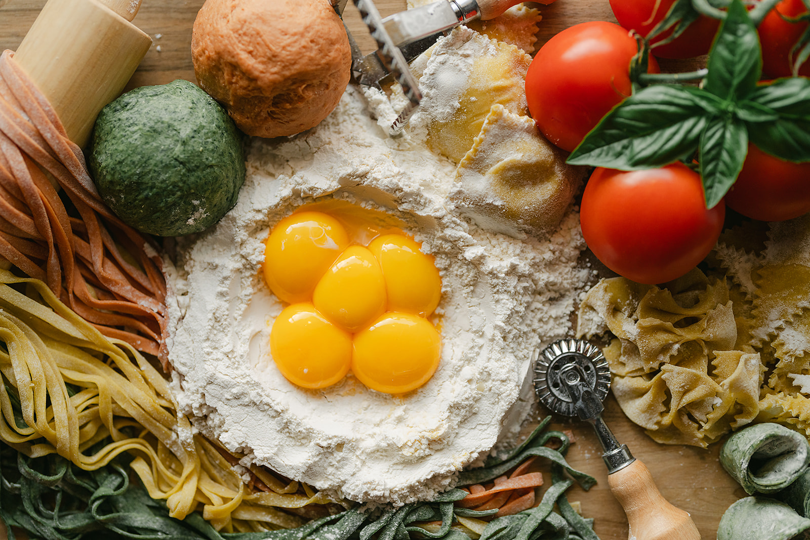 Ingredients for making pasta lay flat on a table. Raw eggs nestled in flour are surrounded by fresh pasta dough, already made pasta in various shapes, fresh tomatoes, and a pasta cutter.